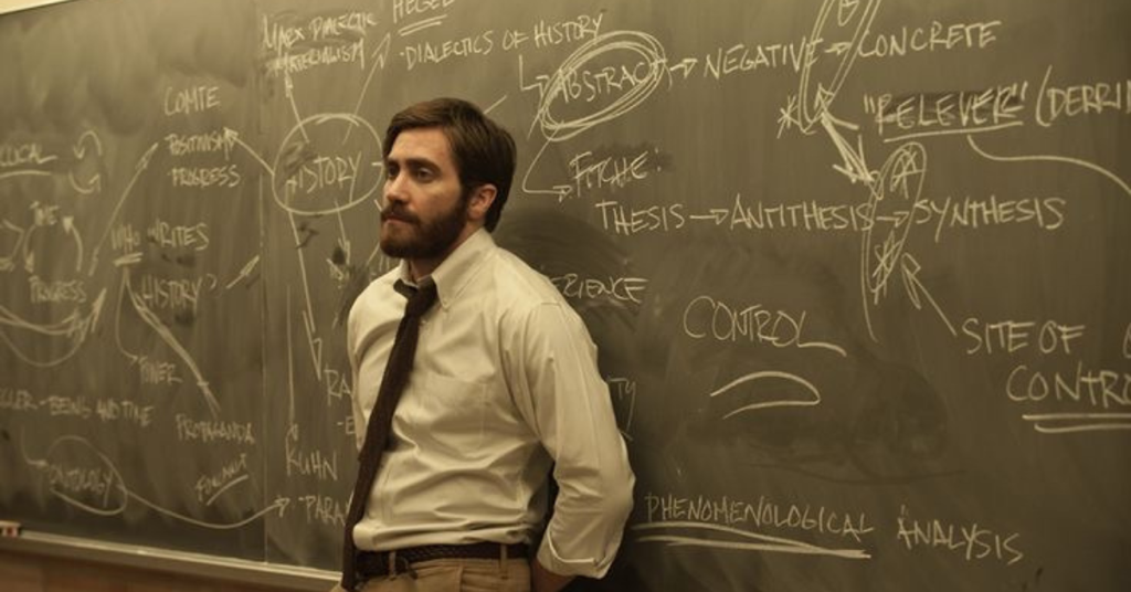Professor leaning against chalk board with chalk writing on it.